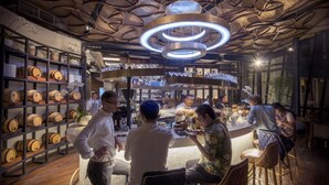 Eine Bar mit Holzfässern in Regalen, einer runden Deckenleuchte und Gästen, die sitzen und stehen.