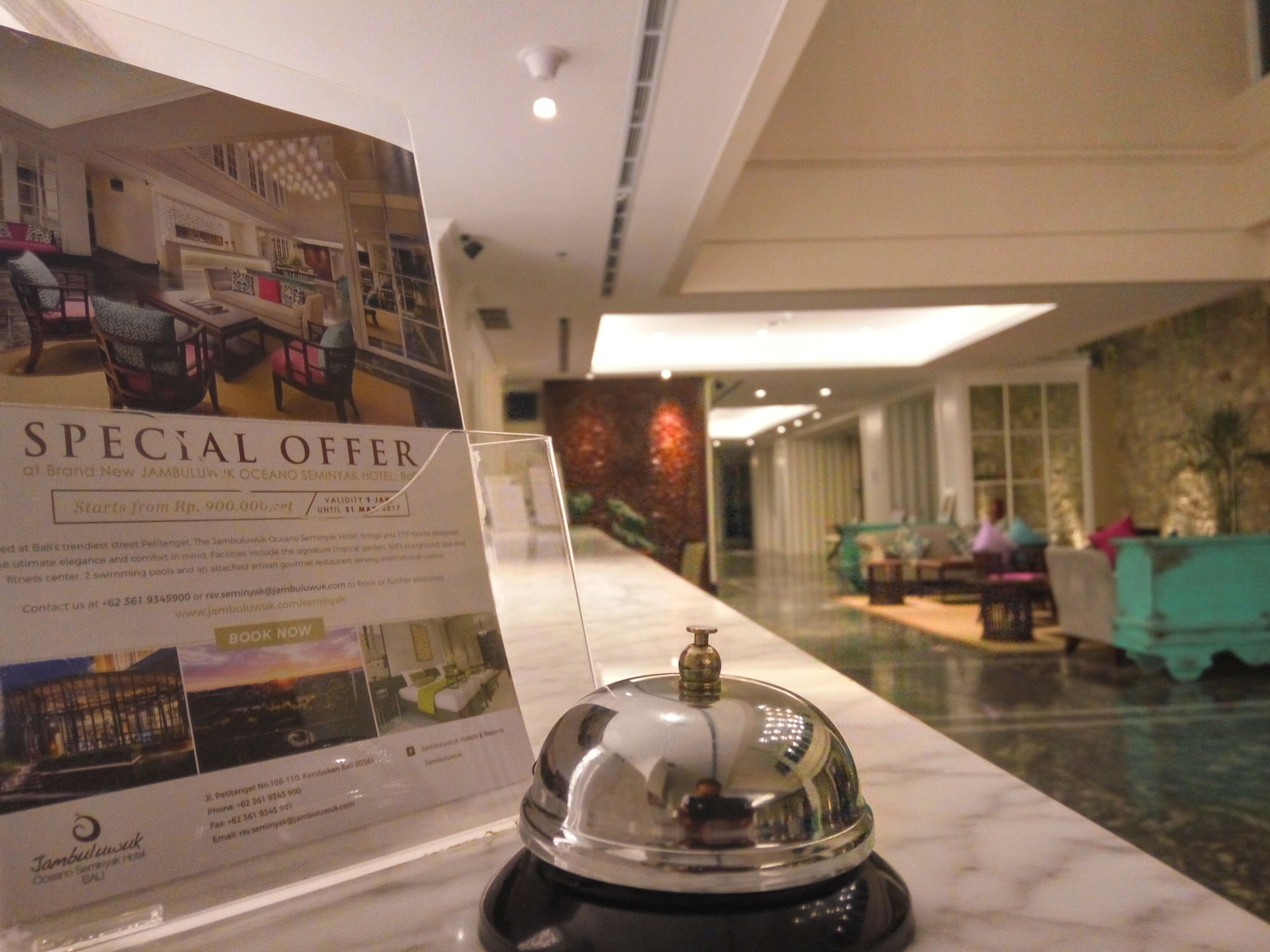 Un espace d'accueil d'hôtel avec une pancarte annonçant une offre spéciale, un comptoir de réception et un aperçu du hall d'entrée, avec des sièges et un mur décoratif.