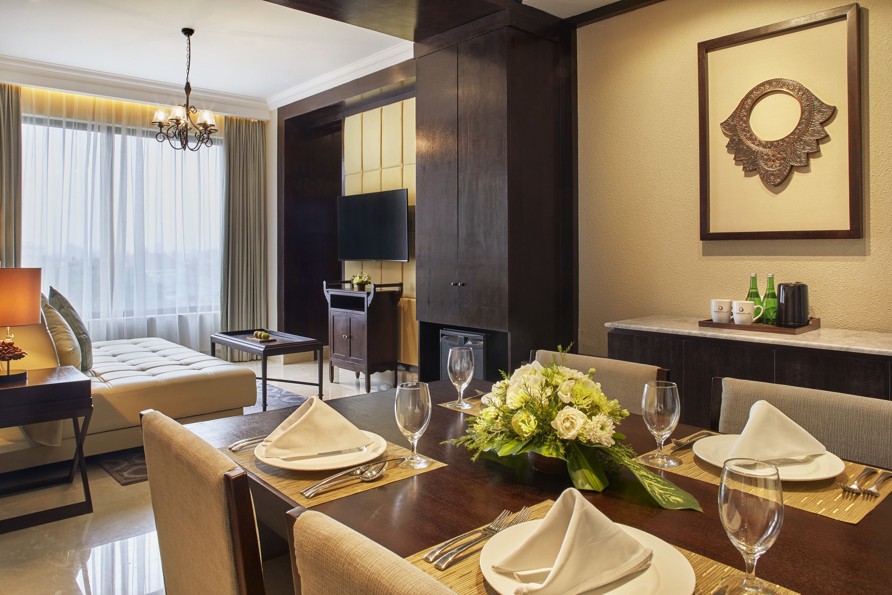 A hotel dining area with a wooden table set for a meal, a sofa, and a television in the background.
