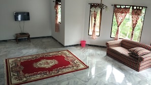 A living room with a red and beige patterned carpet, a plaid sofa, a television on a stand, and a window with curtains. - Villa Sabrina Bumi Ciherang (Cipanas)