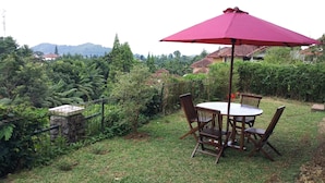 A round wooden table with four chairs and a red umbrella, surrounded by lush greenery and a stone wall. - Villa Tamie Bumi Ciherang (Cipanas)