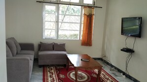 A living room with a sofa, a coffee table, a TV mounted on the wall, and a window with curtains. - Villa Tamie Bumi Ciherang (Cipanas)