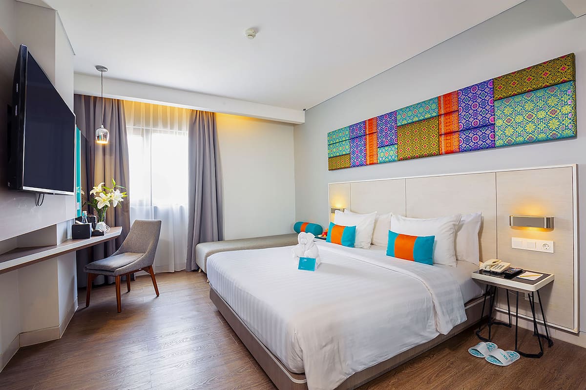 A hotel room with a large bed, a desk, a chair, a TV, and a colorful wall art piece.