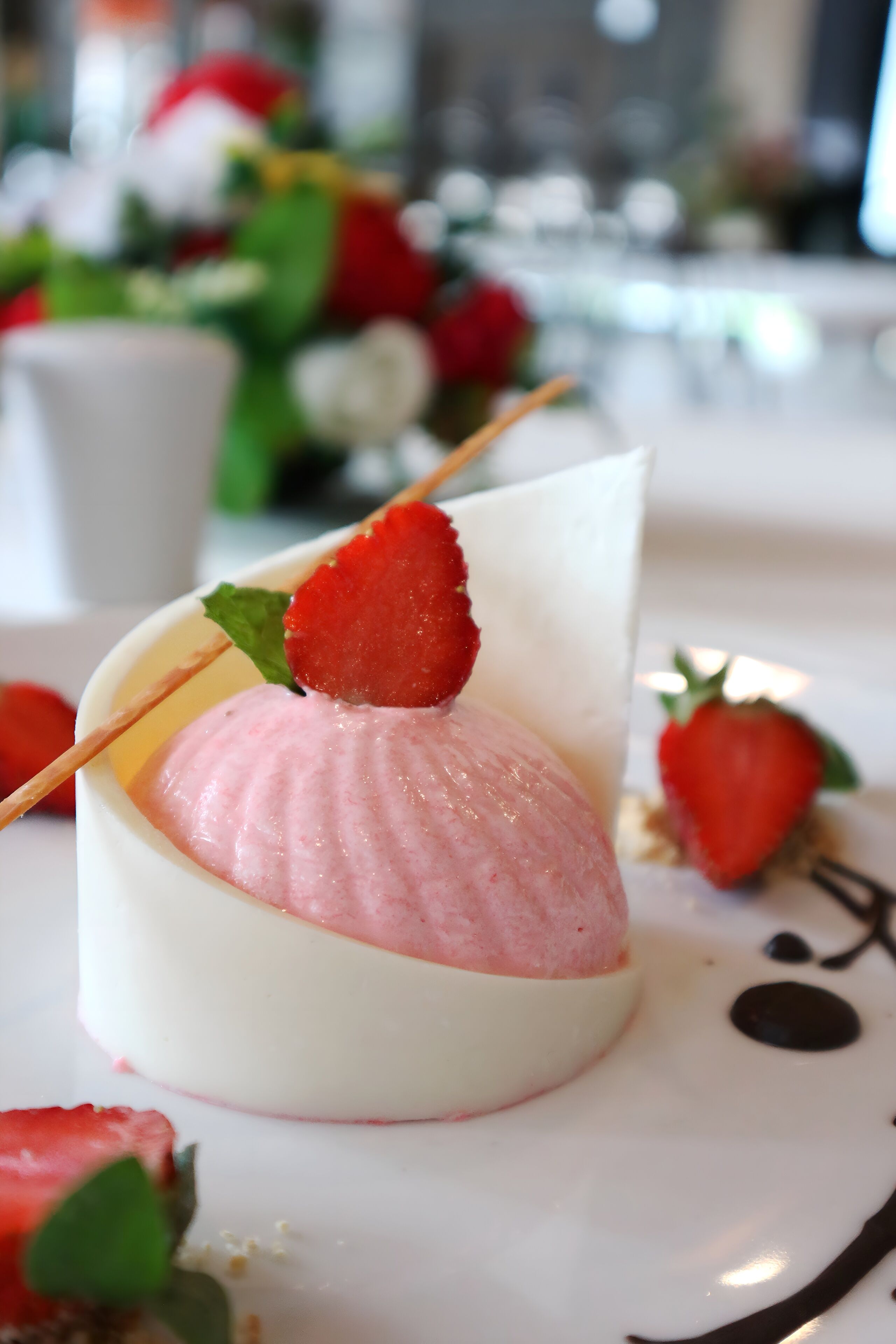Un dessert composé d’une fraise enfilée sur une brochette, présenté sur une assiette blanche ornée d’un motif en chocolat.