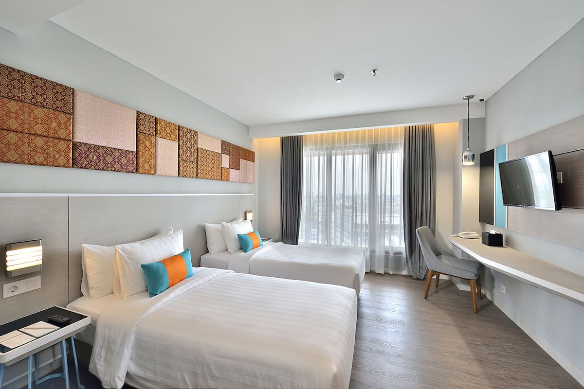 A hotel room with two beds, a desk, a chair, a TV, and a large window with curtains.