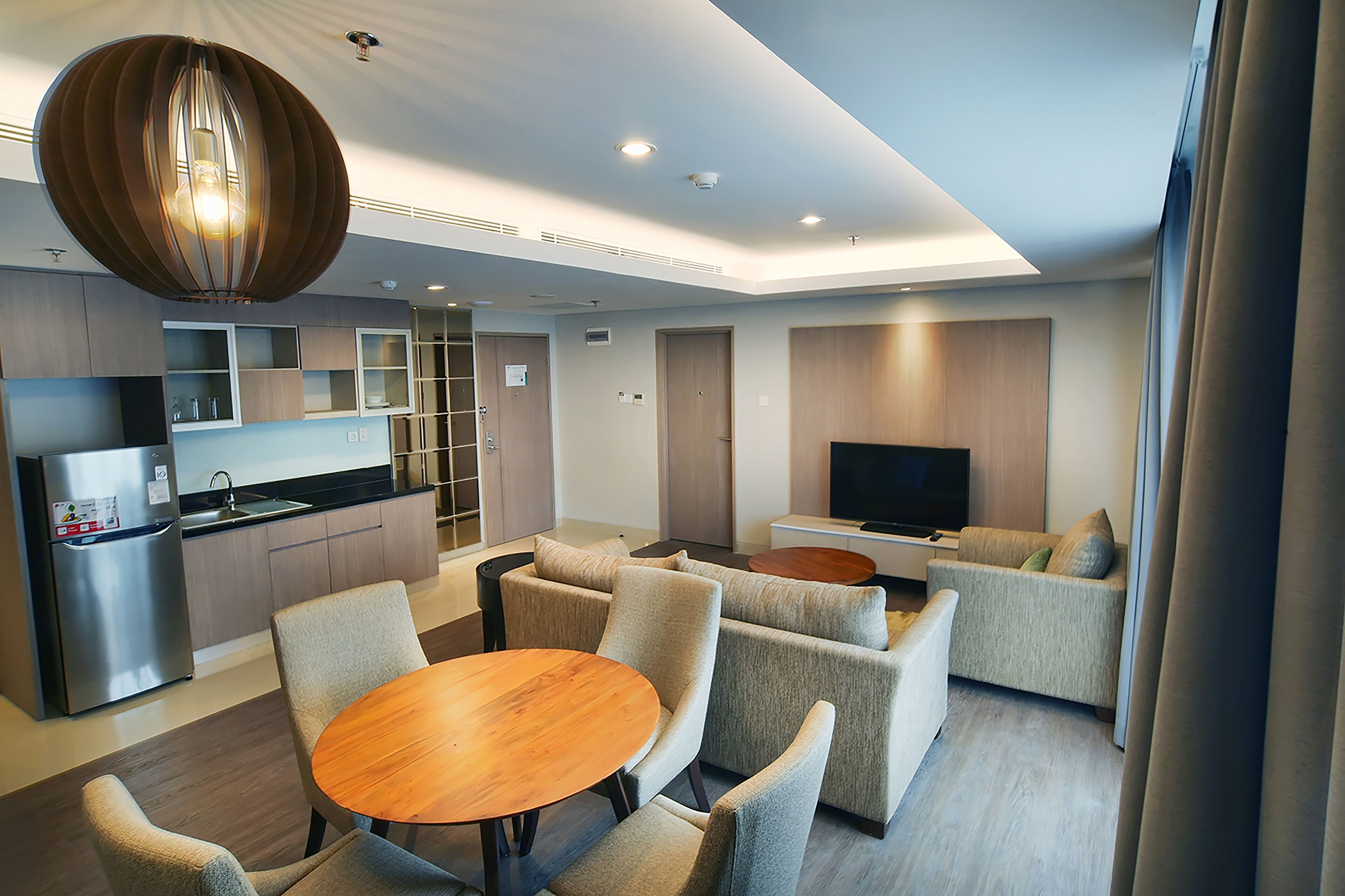 A modern hotel room with a kitchen area, a dining table with chairs, a sofa, and a TV.