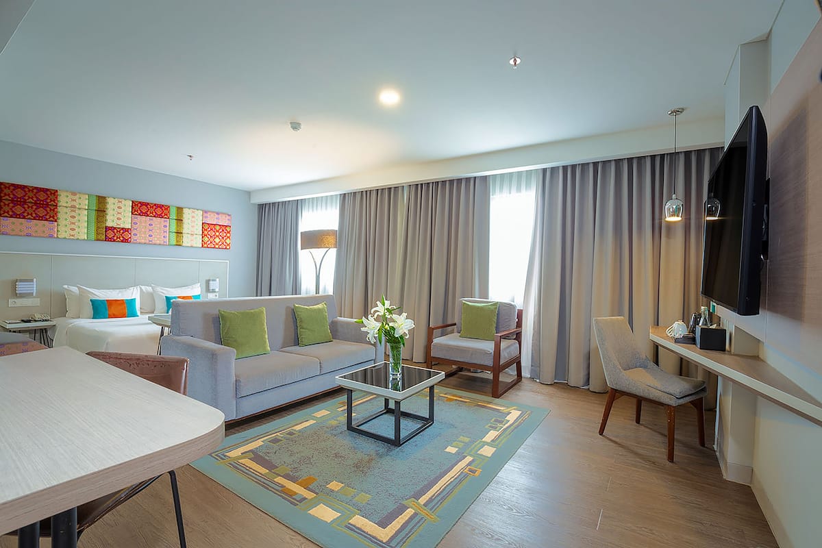 A modern hotel room with a sofa, armchairs, a coffee table, a TV, and a large window with curtains.
