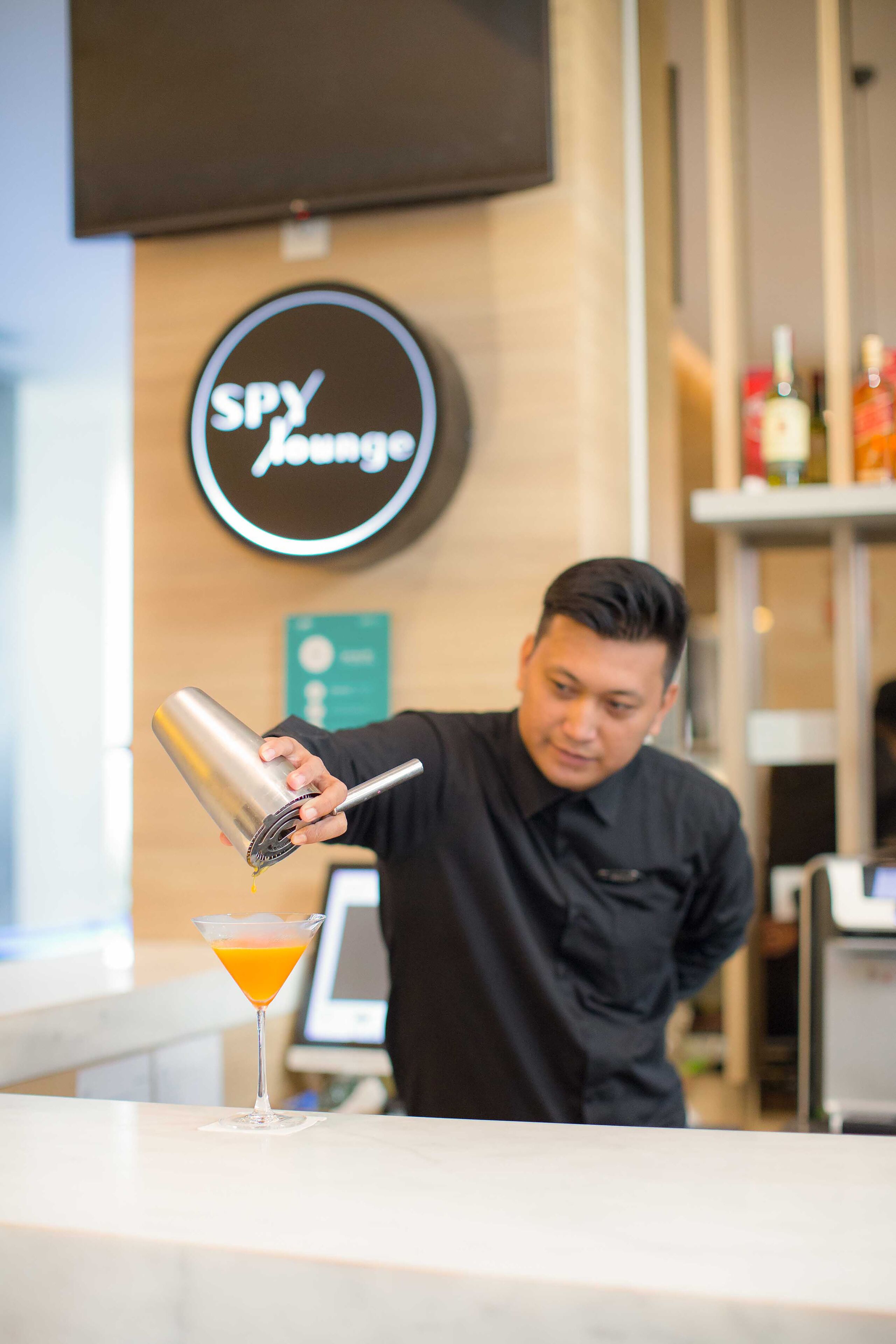 A bartender preparing a cocktail at a bar with a sign reading "SPY Lounge" in the background.