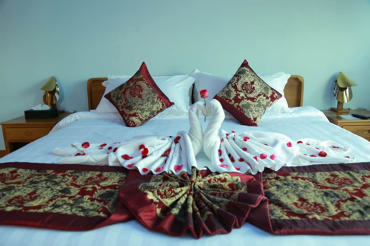 A neatly made bed with a white and red blanket, two decorative pillows, and a rose on the pillow.