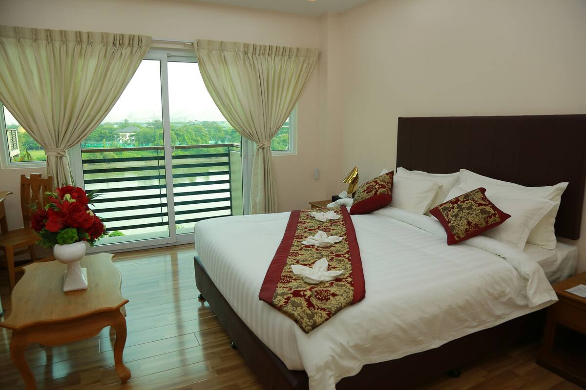 A hotel room with a large bed, a wooden table with a vase of flowers, and a view of greenery through the window.