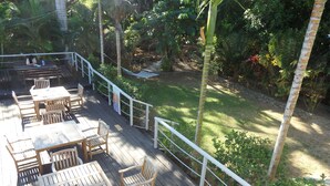 Une terrasse en bois, agrémentée de tables et de chaises, entourée d’une balustrade blanche, et offrant une vue sur un jardin où poussent différentes plantes et arbres.