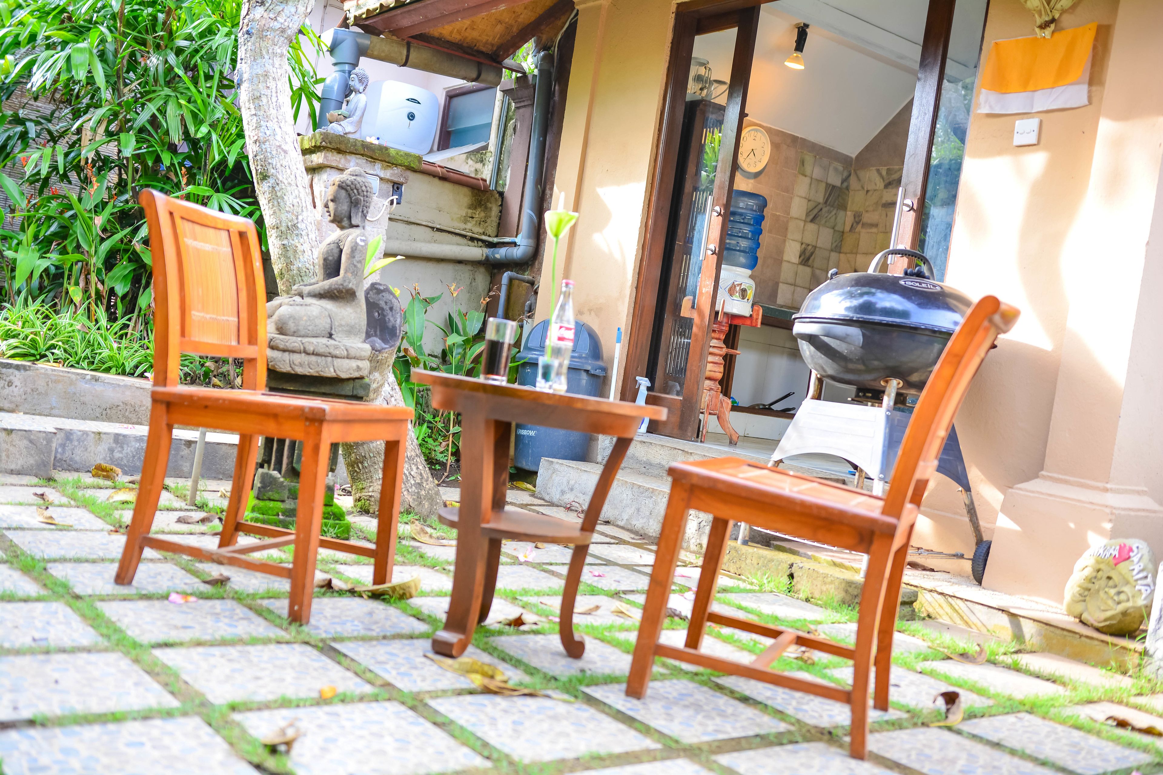An outdoor seating area with wooden chairs and a table, surrounded by greenery and a stone pathway.