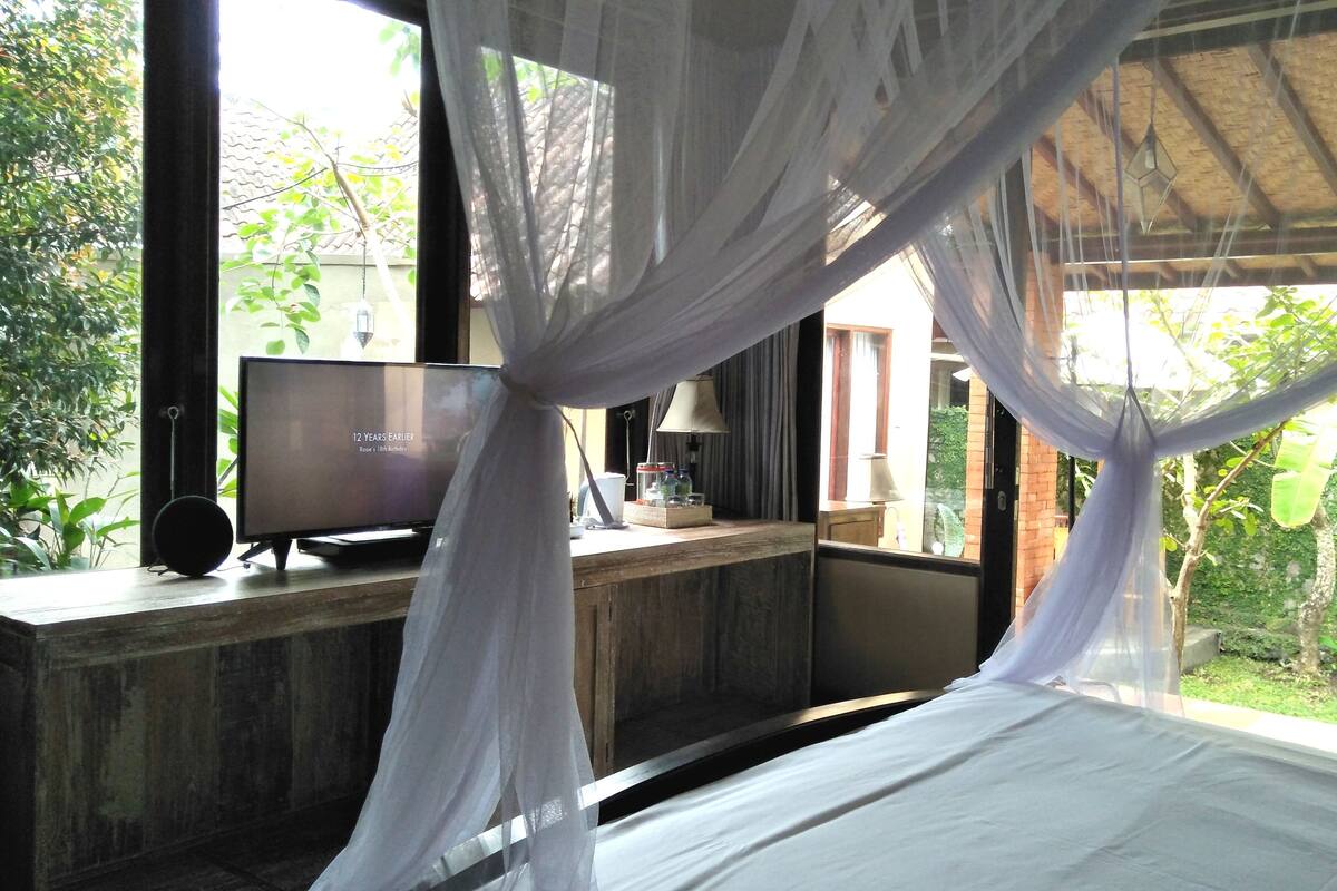 A bedroom with a bed, a TV on a wooden desk, and a view of greenery outside.