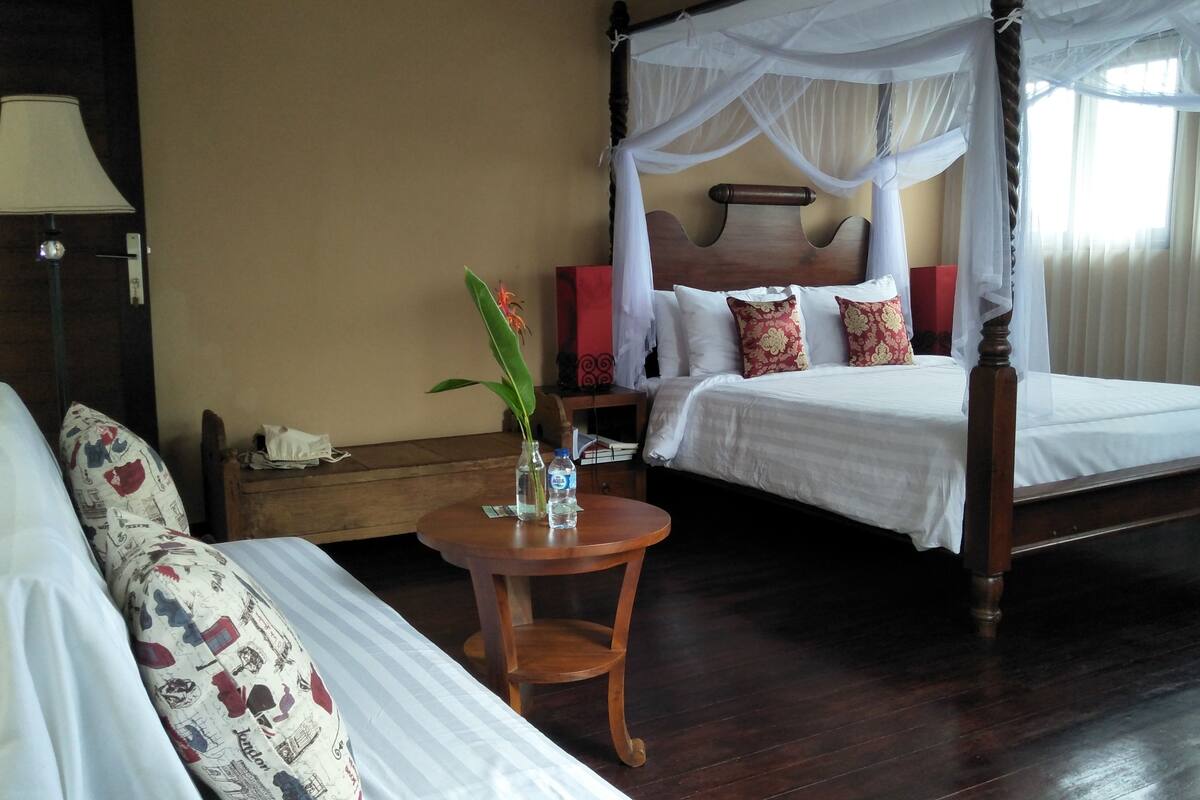 A hotel room with a four-poster canopy bed, a bedside table with a vase and water bottles, a sofa with patterned cushions, and a nightstand with a lamp.