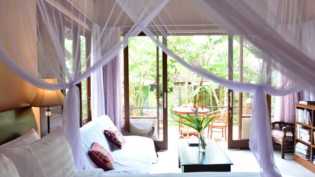 A bedroom with a bed, bedside table, and a view of the garden through large windows. - Griya Sriwedari (Ubud)