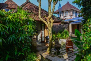 A courtyard with a tiled floor, a wooden chair, a statue, and a building with a tiled roof. - Griya Sriwedari (Ubud)