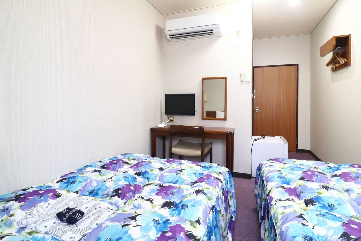 Quarto de hotel com duas camas, uma escrivaninha, uma cadeira, uma televisão, um espelho e um aparelho de ar condicionado.