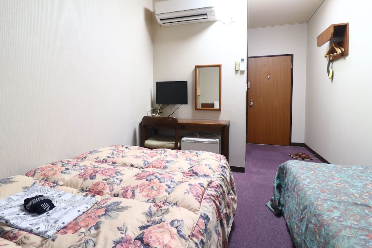 Quarto de hotel com cama, escrivaninha, cadeira, ar condicionado, espelho e porta.