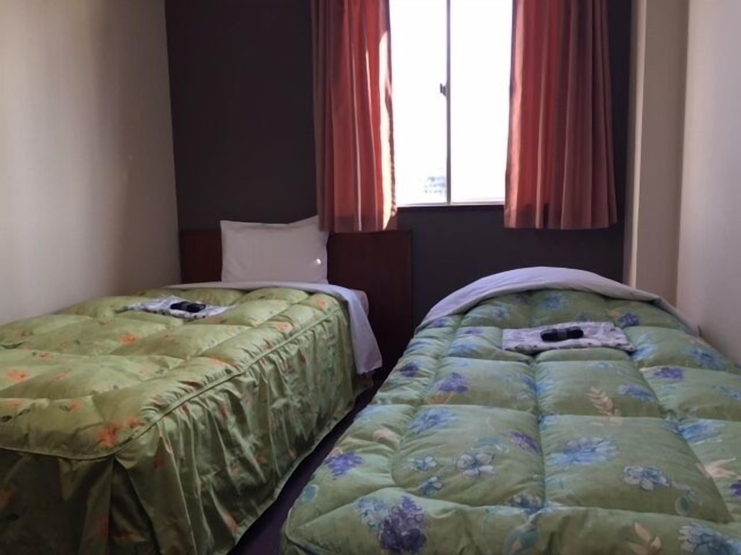 A room with two single beds, each with a floral duvet cover. There is a window with red curtains, and the room has a simple, functional design.