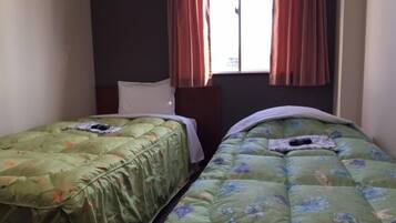 A room with two single beds, each with a floral duvet cover. There is a window with red curtains, and the room has a simple, functional design.