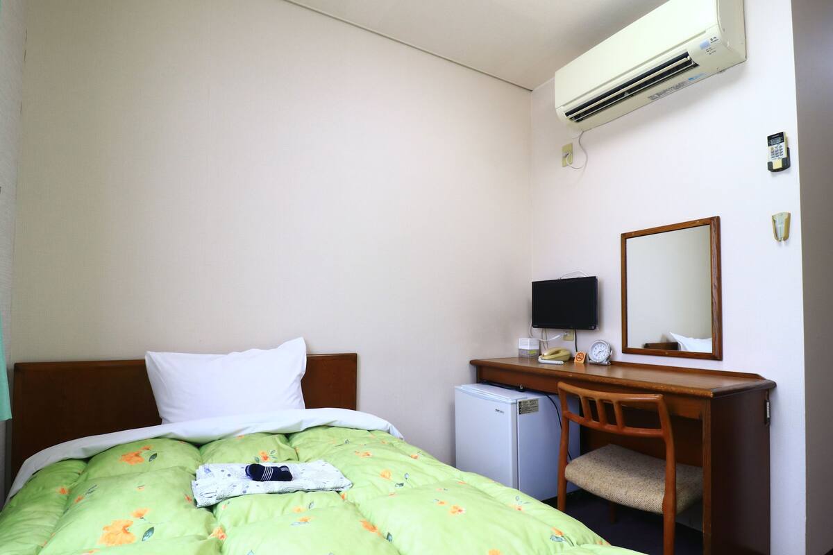 Quarto de hotel com cama, escrivaninha, cadeira, frigorífico pequeno, ar condicionado, televisão e espelho.