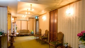 Un hall de hotel con piso alfombrado, sillas de madera, un sofá, una mesita con un jarrón, y una lámpara fijada a la pared.