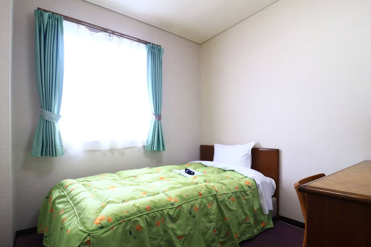 Quarto de hotel pequeno com uma cama de solteiro, uma mesinha de cabeceira de madeira e uma janela com cortinas.