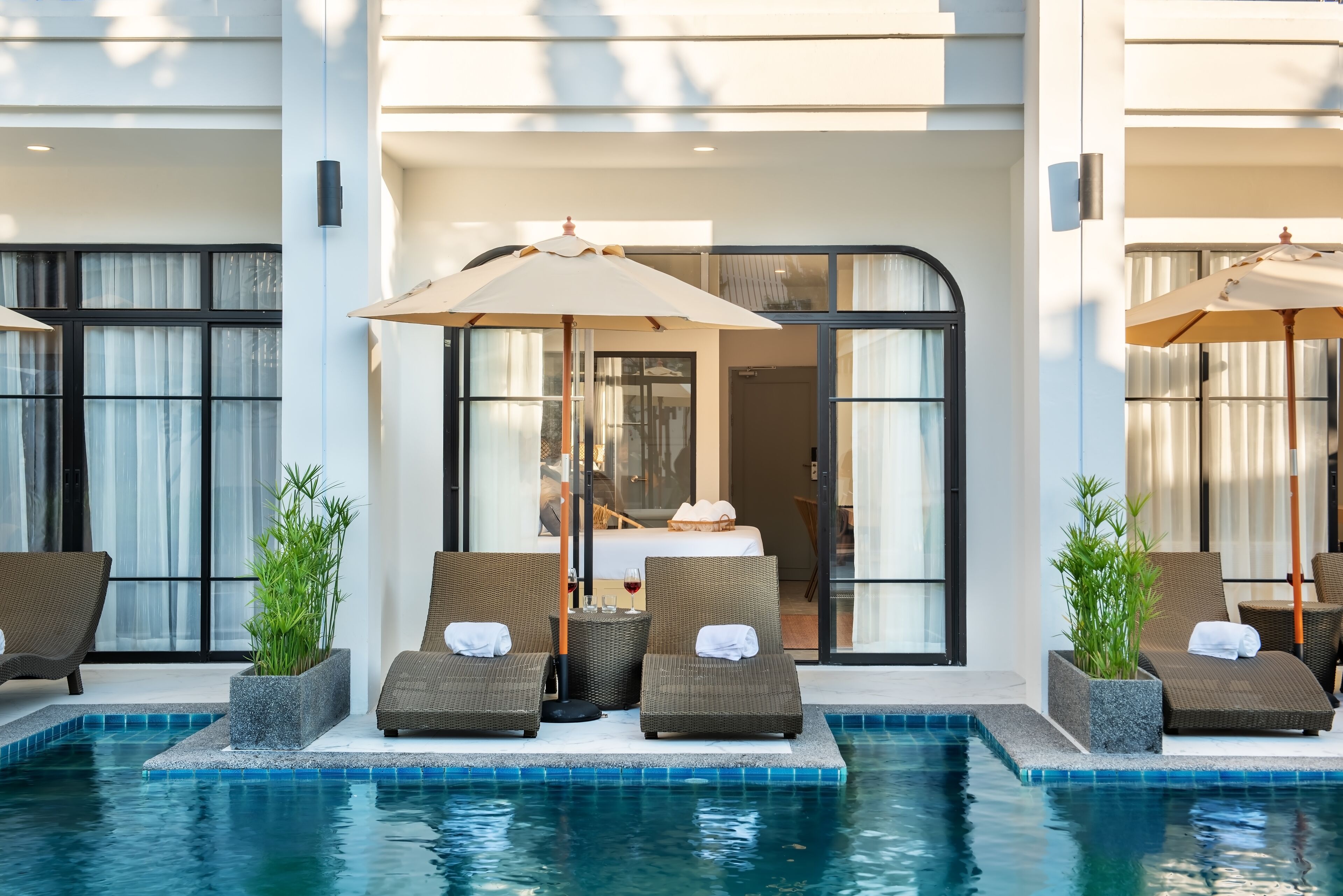 Zona de piscina con sillones de descanso y sombrillas, entrada con puerta de cristal y vista al interior.