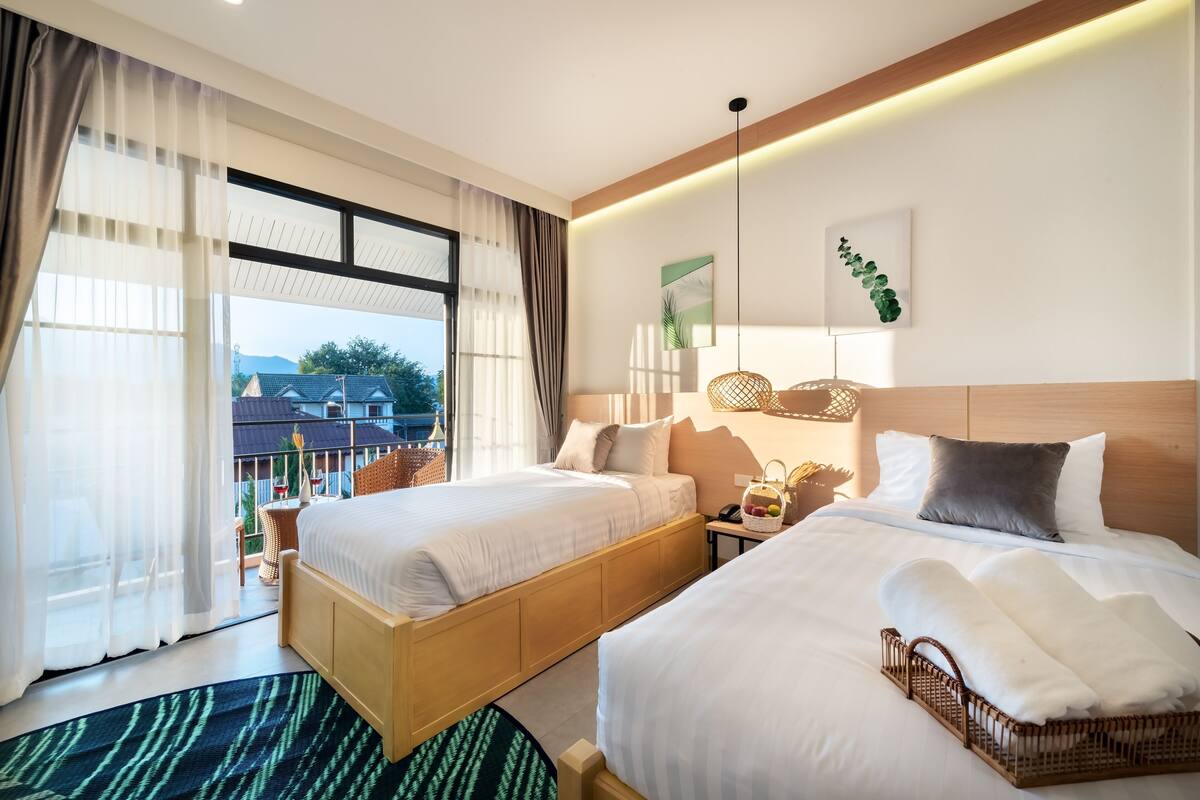 Habitación de hotel con dos camas, un ventanal con cortinas, una mesita de noche con lámpara y vistas a edificios.