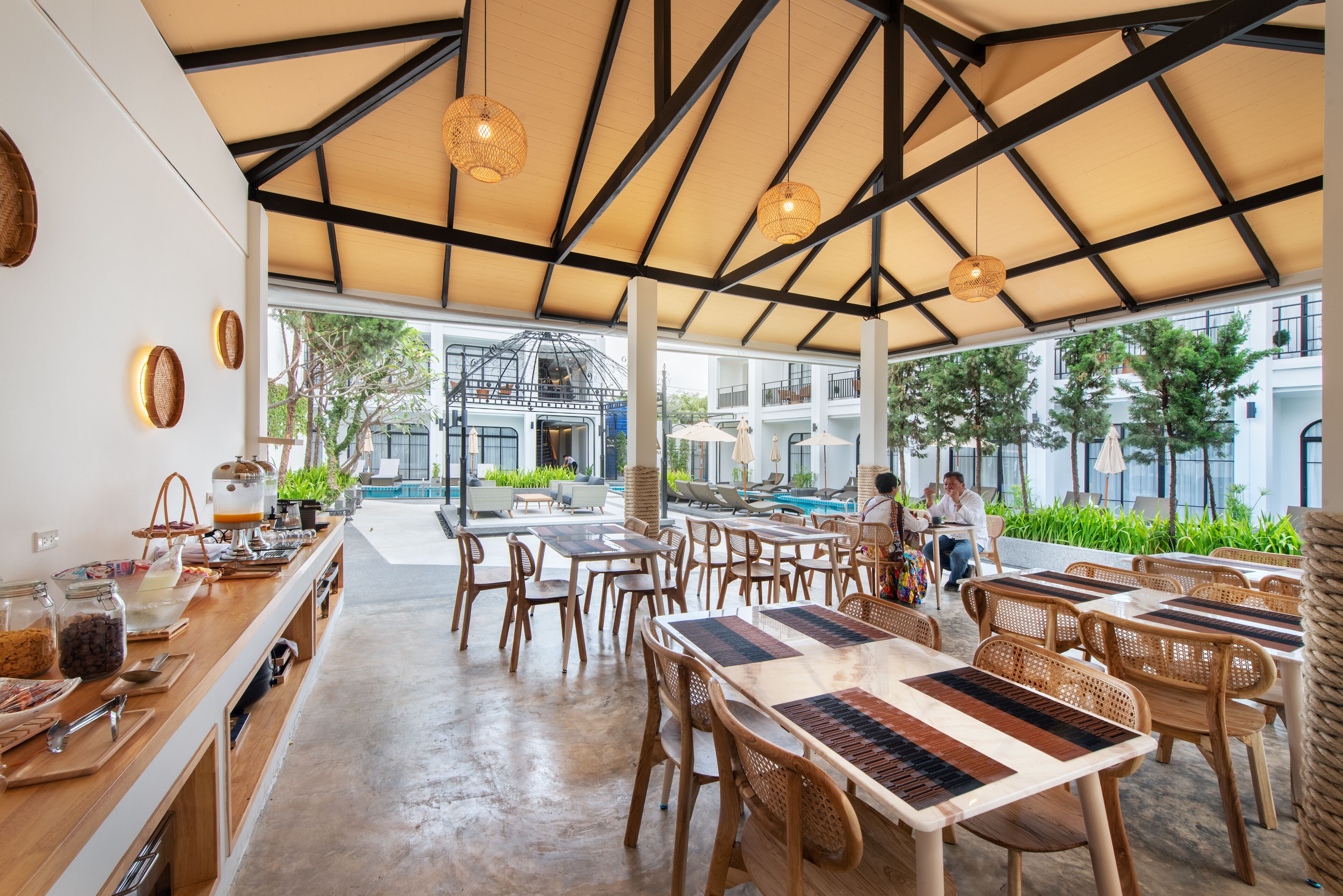 A spacious outdoor dining area with wooden tables and chairs, a buffet counter with food items, and a view of a swimming pool and building in the background.