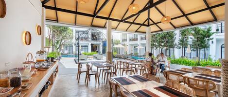 A spacious outdoor dining area with wooden tables and chairs, a buffet counter with food items, and a view of a swimming pool and building in the background.