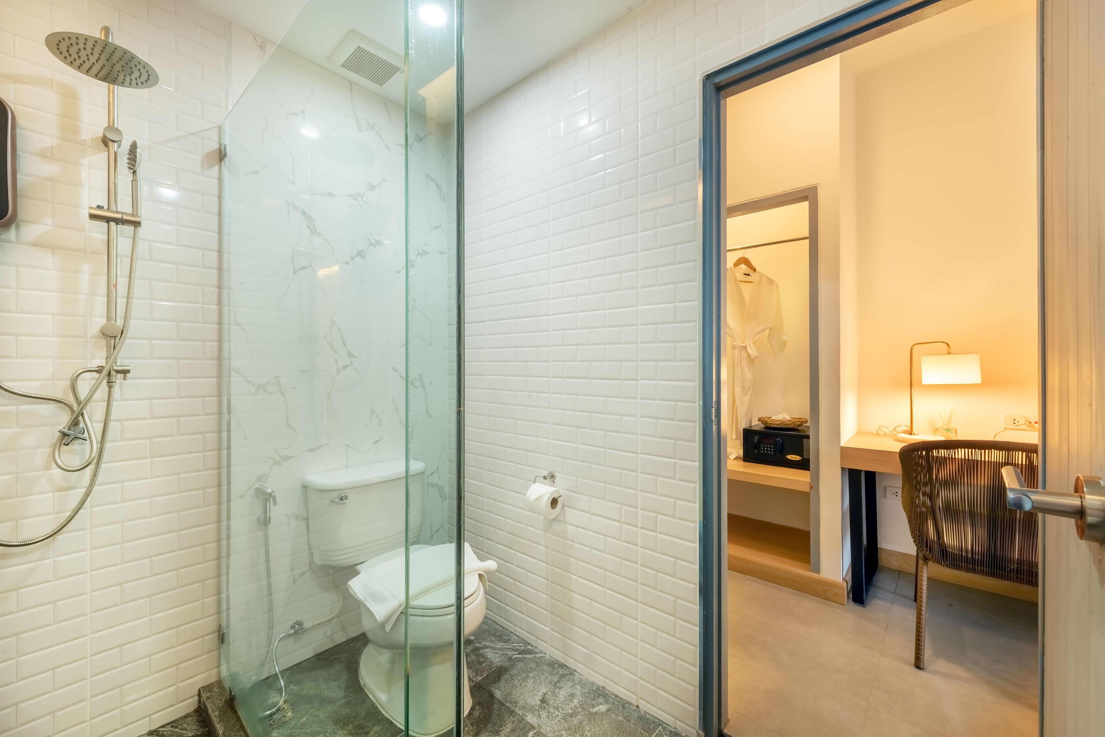 Un baño moderno con ducha de vidrio, paredes con azulejos blancos, inodoro y tocador con silla.