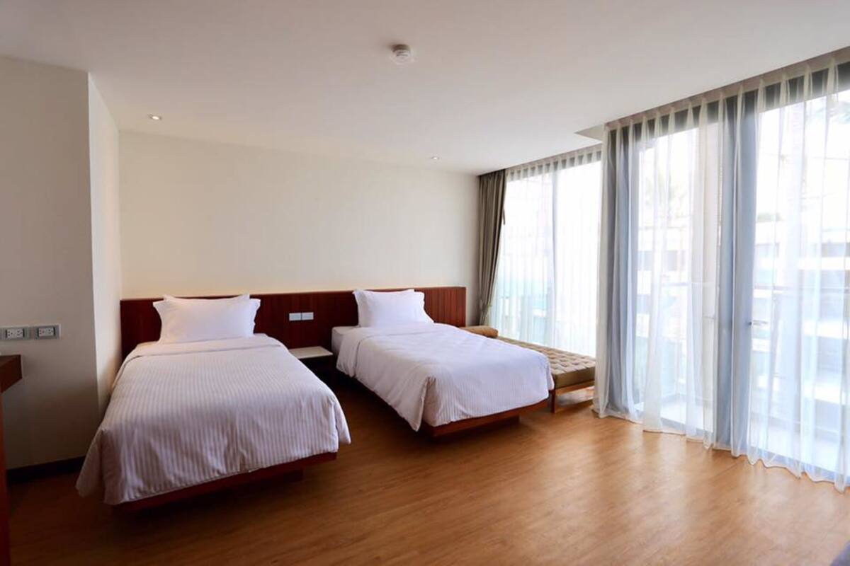 A hotel room with two beds, wooden flooring, and large windows with sheer curtains.