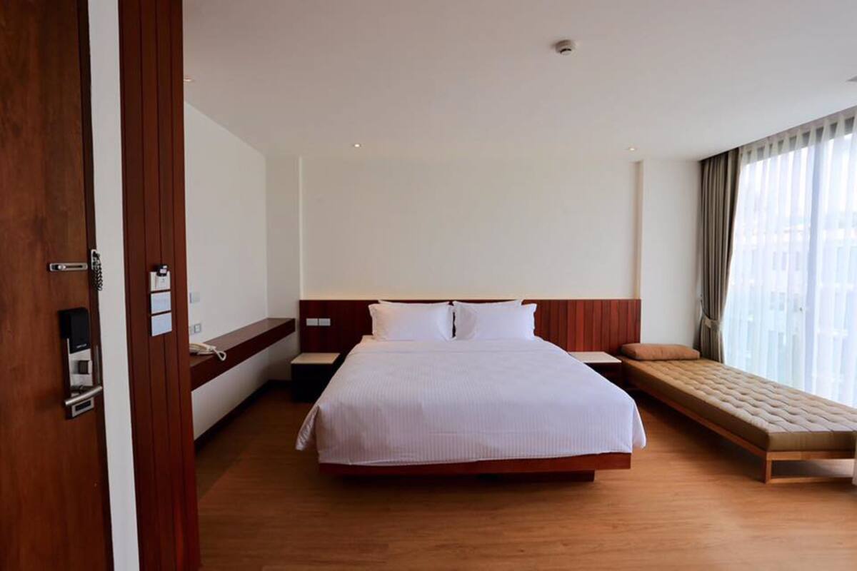 A hotel room with a large bed, a bedside table, a bench, a window with curtains, and a wooden floor.