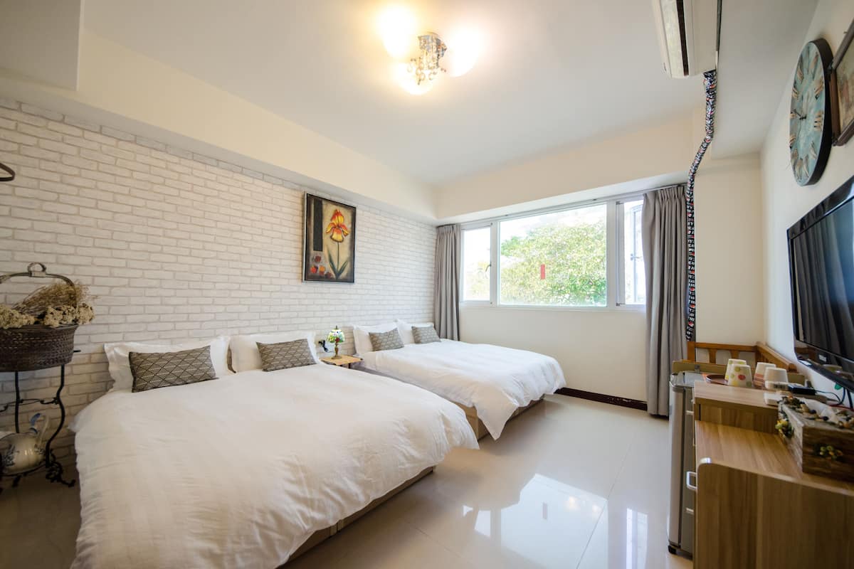 A hotel room with two beds, a large window, a TV, and a brick wall.