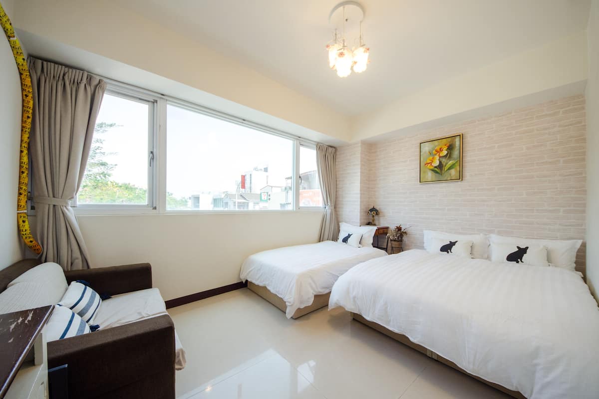 A hotel room with two beds, a desk, a chair, a window with curtains, a brick wall, and a chandelier.