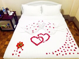 Una cama bien tendida con sábanas blancas, una decoración con forma de corazón hecha de pétalos de rosa y un ramo de rosas rojas.