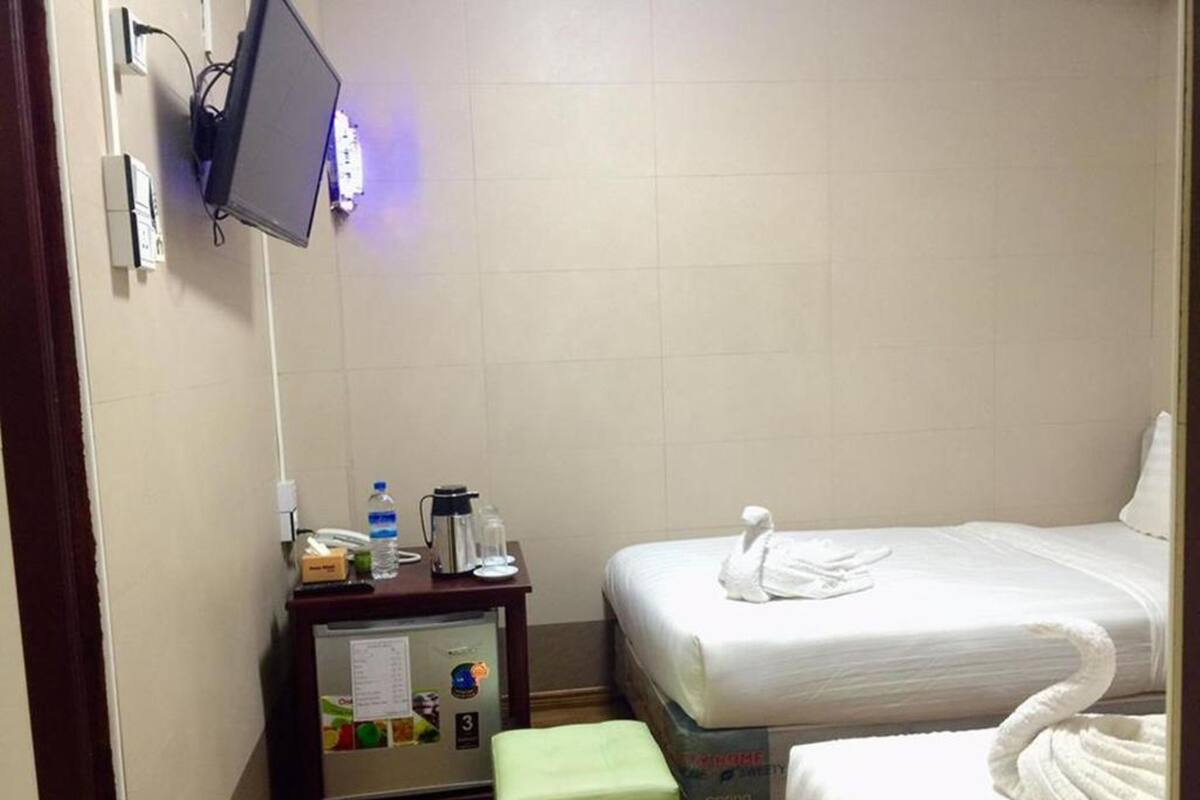 A hotel room with a bed, a small table with a kettle, a water bottle, and a refrigerator.
