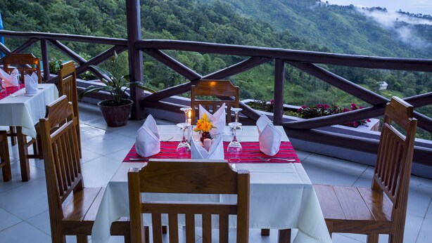 Un área de comedor al aire libre con mesas dispuestas para dos, sillas de madera y vistas a colinas verdes exuberantes.