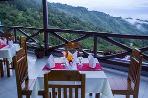 An outdoor dining area with tables set for two, wooden chairs, and a view of lush green hills. - The Floral Breeze Hotel Mt.Victoria (Kanpetlet)