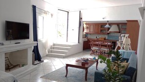 Una sala de estar con televisor, chimenea, comedor con sillas, cocina con armarios y una escalera que sube a un piso superior.