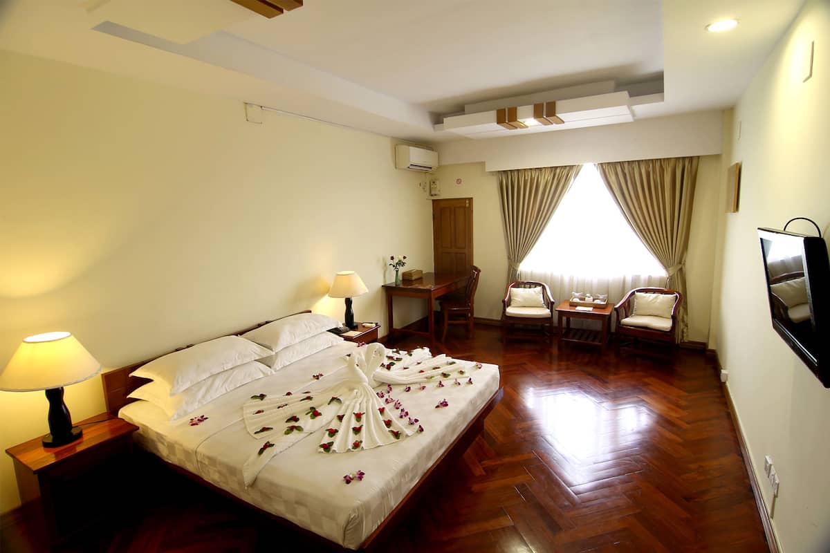 Une chambre d’hôtel avec un grand lit, deux tables de chevet avec des lampes, un plancher en bois, un bureau avec une chaise, une télévision et une fenêtre avec des rideaux.