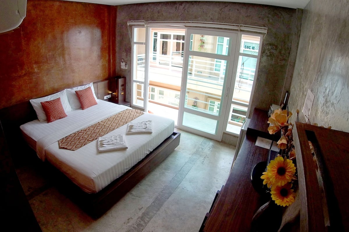 A hotel room with a bed, a desk with a vase of flowers, and a view of the outside through a large window.