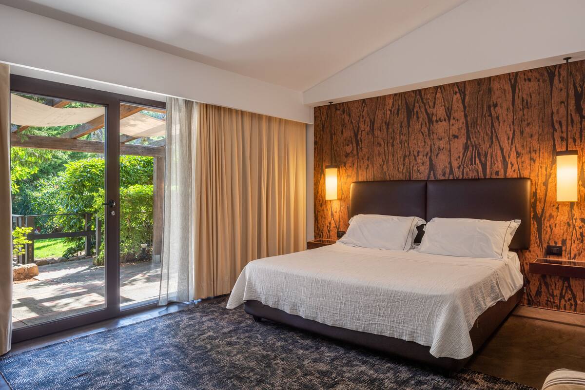 A hotel room with a large bed, a wooden accent wall, and a sliding glass door leading to a balcony with greenery.