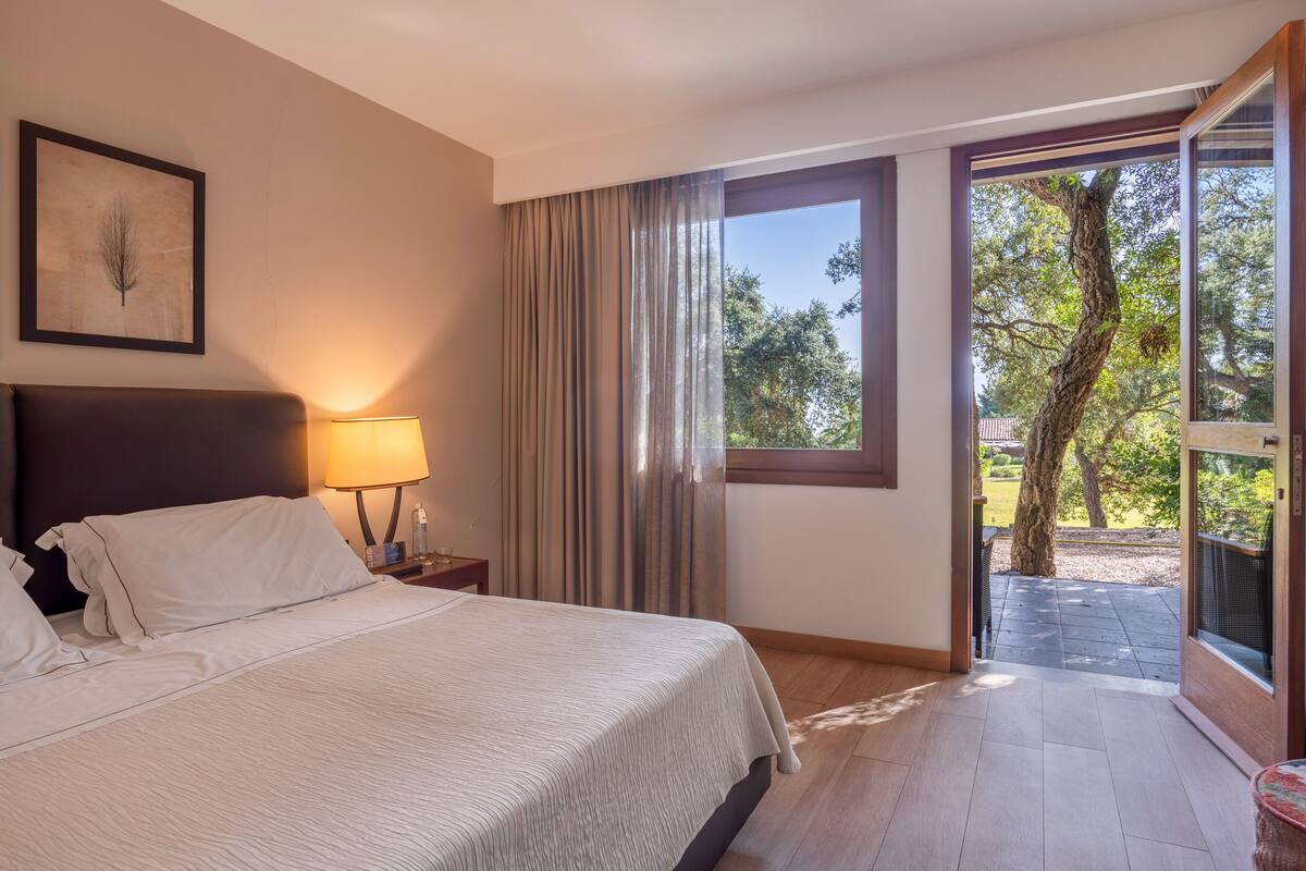A bedroom with a bed, a nightstand, a lamp, a window with curtains, and a view of the outdoors.
