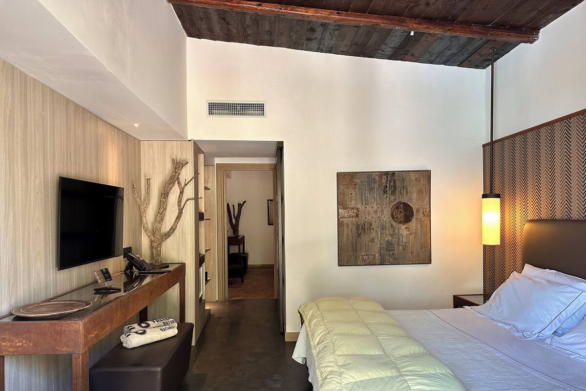 A modern hotel room with a flat-screen TV, a wooden desk, a bed with a green duvet, and a wall-mounted artwork.