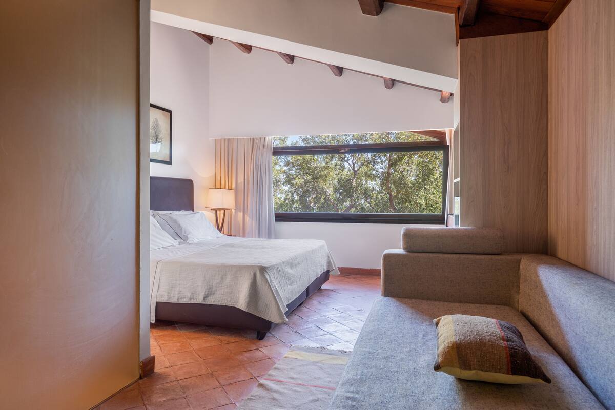 A bedroom with a bed, a sofa, a window with a view of trees, and a lamp.