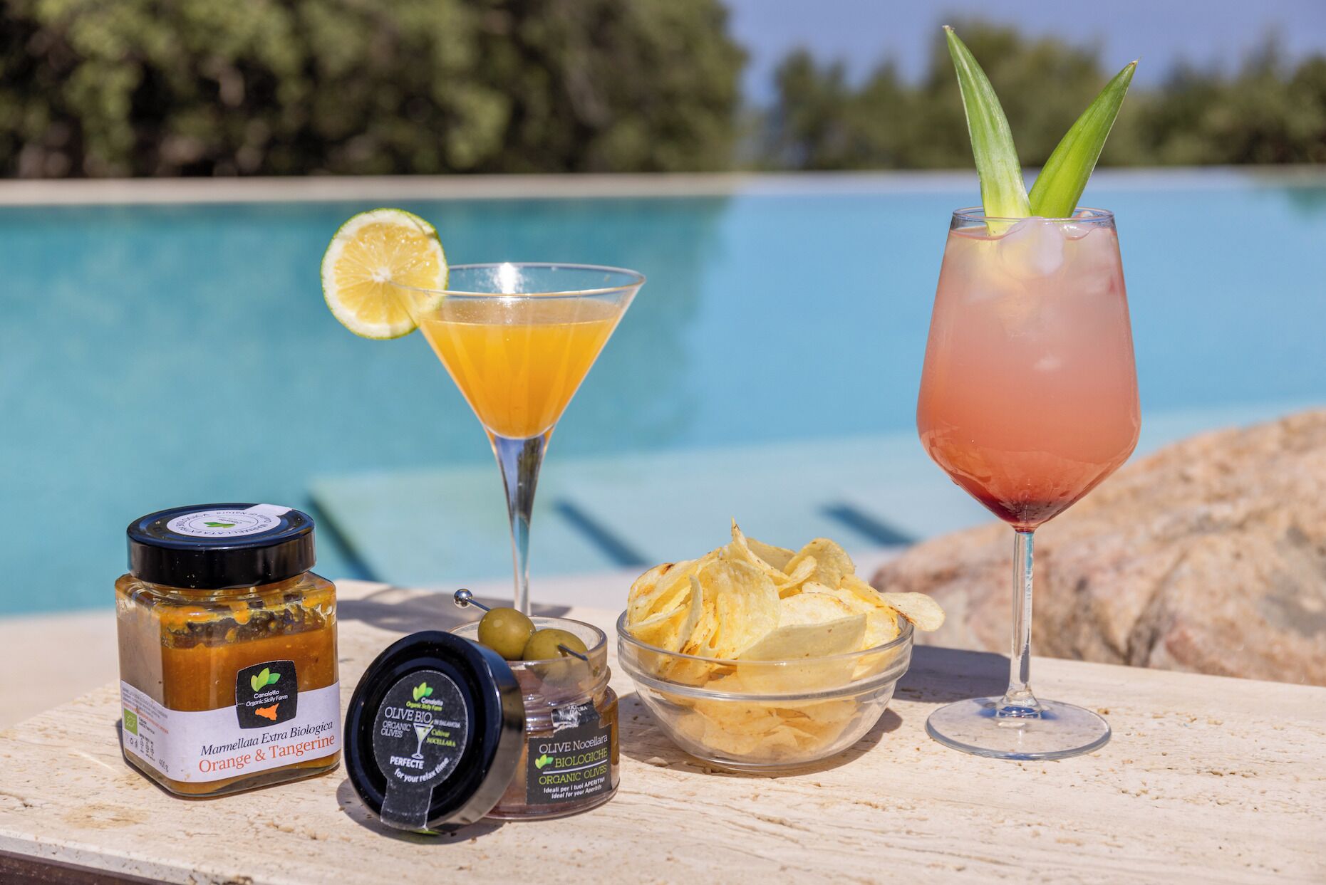Una mesa con una botella de aceite de oliva orgánico, un frasco de aceite de oliva, un bol de patatas fritas y dos bebidas, con una piscina al fondo.