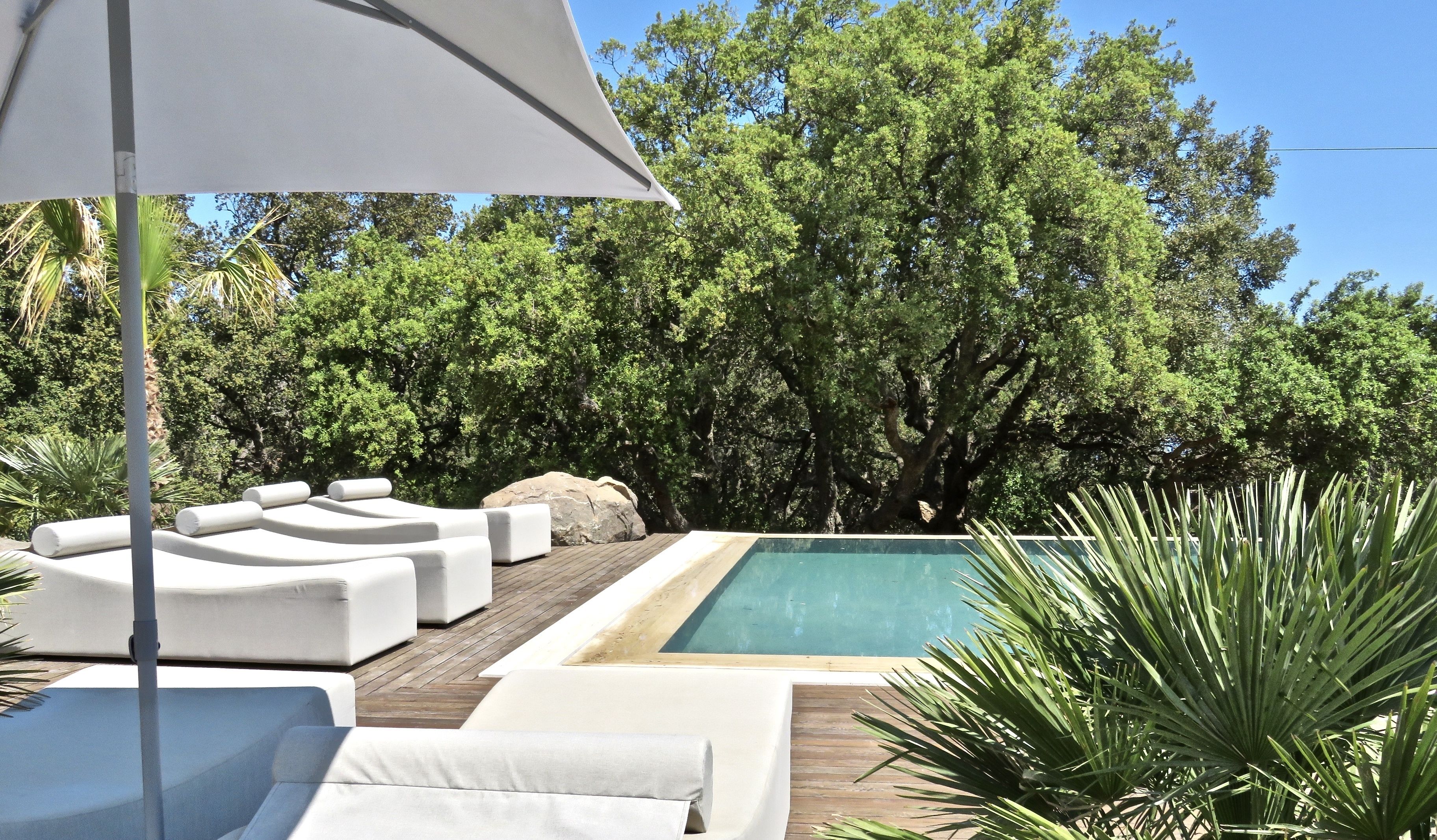 Zona junto a la piscina con sillas reclinables blancas y una sombrilla grande.
