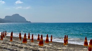 A beach with rows of sun loungers and umbrellas, a rocky shoreline, and a distant mountain. - Vallegrande Nature Resorts (Cefalù)